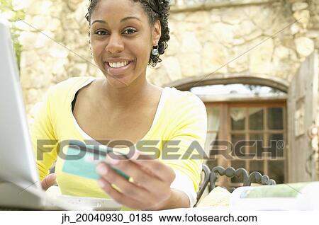 Young woman sitting at a table in front of a laptop holding a credit card smiling View Large Photo Image Stock Photography - Young woman sitting at a table in front of a laptop holding a credit card smiling. Fotosearch