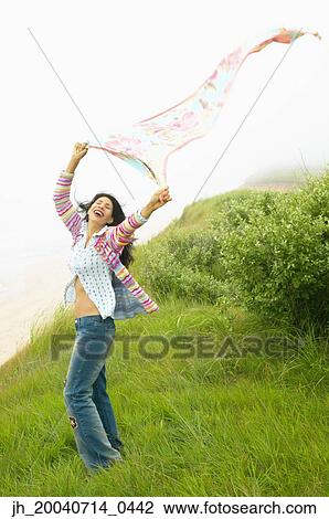Stock Image - Young woman standing in a field holding a scarf above her head. Fotosearch