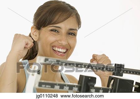 Stock Photo - Young woman standing on a weighing scale with arms raised in excitement. Fotosearch