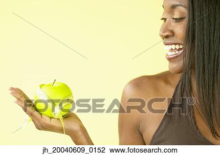 Stock Image - Mid adult woman smiling holding an inch tape around an apple. Fotosearch