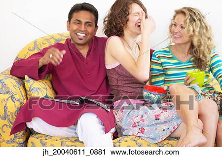 Stock Image - Two young women laughing sitting on a couch with a young man. Fotosearch