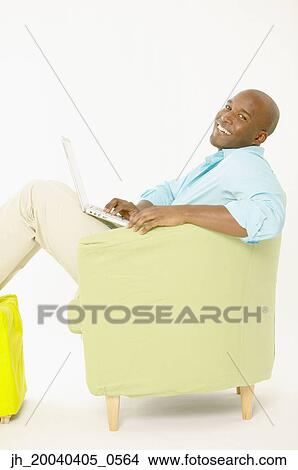 Young man sitting on a couch operating a laptop View Large Photo Image Picture - Young man sitting on a couch operating a laptop. Fotosearch