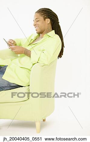 Young man sitting on a couch operating a hand held device View Large Photo Image Stock Image - Young man sitting on a couch operating a hand held device. Fotosearch
