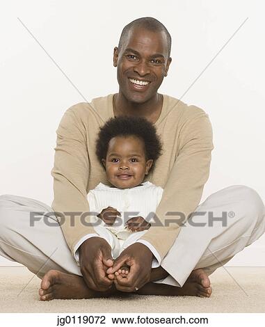 Stock Image - Father sitting cross legged while holding baby daughter. Fotosearch