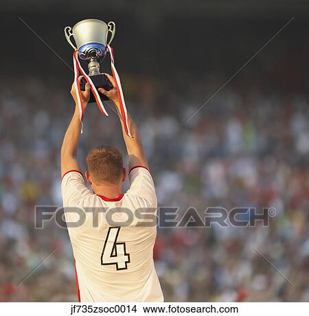 Picture - Male soccer player triumphantly holding up trophy. Fotosearch