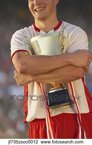 Stock Image - Male soccer player triumphantly hugging trophy. Fotosearch