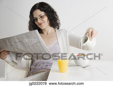 Stock Photo - Woman pouring milk on cereal while reading newspaper. Fotosearch