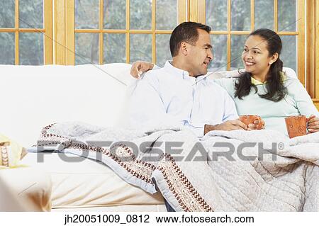 Stock Image - Couple sitting on the couch under a blanket together. Fotosearch