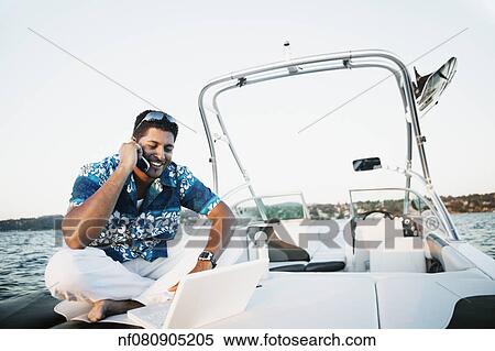 Young man talking on his cell phone and using a laptop on a boat View Large Photo Image Stock Photography - Young man talking on his cell phone and using a laptop on a boat. Fotosearch