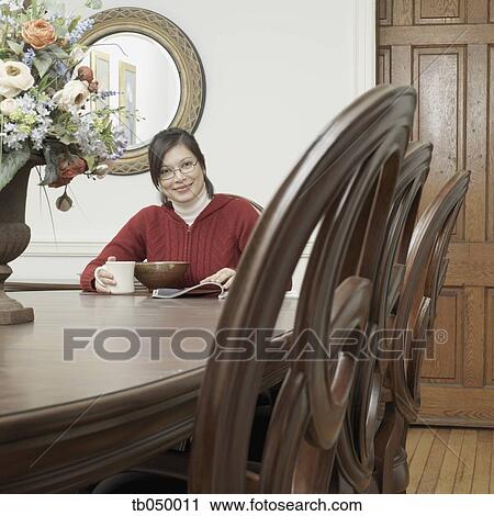 Stock Image - Asian woman eating and reading at dining table. Fotosearch