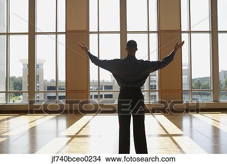 Picture - Businessman standing in sunlit room with arms outstretched, North Bethesda, Maryland, United States. Fotosearch