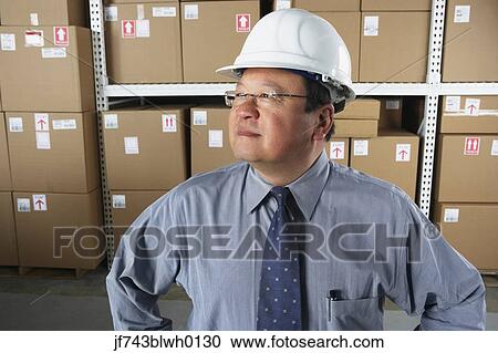Stock Image - Middle-aged Asian businessman wearing hard hat in warehouse. Fotosearch