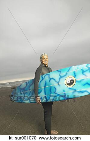 Stock Image - Senior male surfer carrying his surfboard on the beach, Oakland, California, United States. Fotosearch