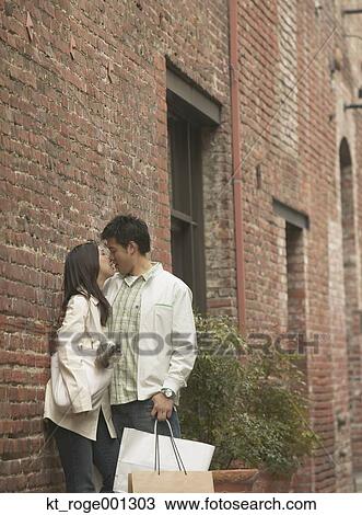 Stock Image - Young Asian couple leaning against a brick building kissing, San Francisco, California, United States. Fotosearch