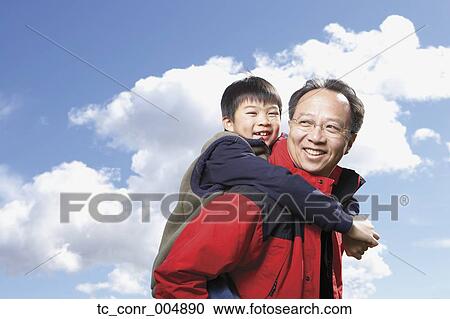 Stock Image - Asian father giving son piggyback ride outdoors. Fotosearch