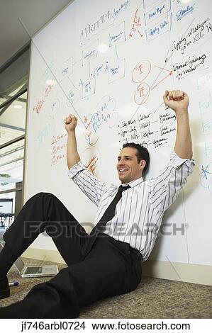 Picture - Businessman sitting on floor cheering in front of whiteboard wall. Fotosearch