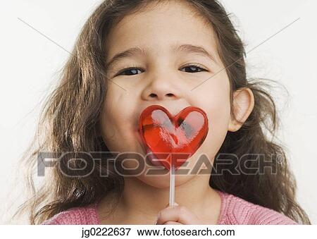 Stock Photo - Studio shot of Hispanic girl licking heart lollipop. Fotosearch