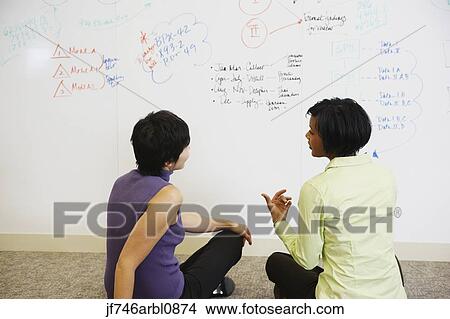 Picture - Two businesswomen sitting on floor in front of whiteboard wall. Fotosearch