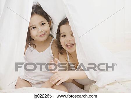 Stock Image - Two young Hispanic sisters playing under sheet. Fotosearch