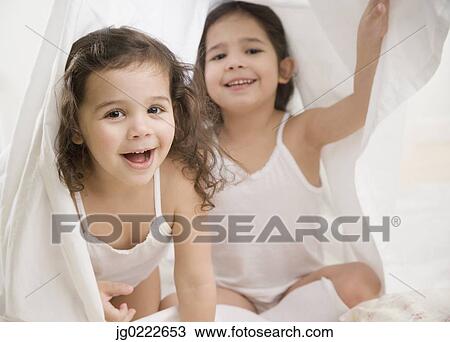 Stock Image - Two young Hispanic sisters playing under sheet. Fotosearch