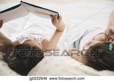 Stock Photo - Two young Hispanic sisters reading a book in bed. Fotosearch