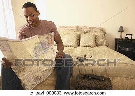 Stock Image - African man sitting on bed looking at map. Fotosearch