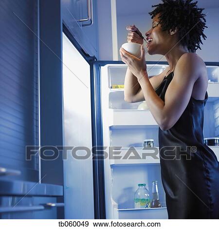 Stock Photo - African woman eating next to open refrigerator. Fotosearch