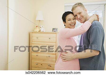Stock Image - Pregnant couple hugging indoors . Fotosearch