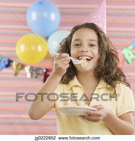 Stock Image - Young girl eating cake at birthday party. Fotosearch