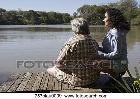 Stock Photo - Couple sitting on wooden dock looking at water. Fotosearch