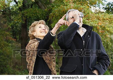 Stock Image - Senior woman pointing while senior man takes photograph outdoors. Fotosearch