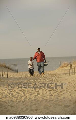 Stock Photograph - African father and son walking on beach with fishing gear. Fotosearch