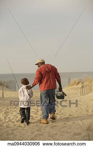 Stock Photograph - African father and son walking on beach with fishing gear. Fotosearch