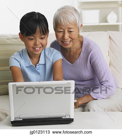 Stock Image - Asian grandmother and granddaughter using laptop on sofa. Fotosearch