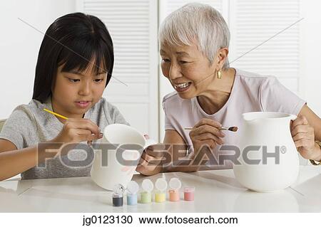 Stock Image - Asian grandmother and granddaughter painting pottery. Fotosearch