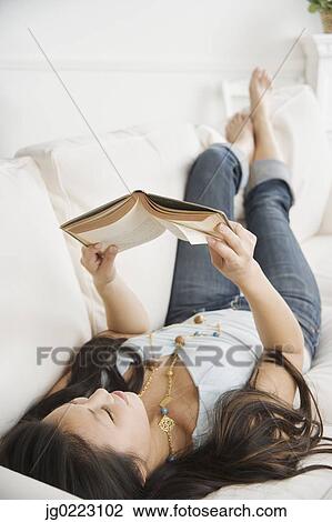 Stock Image - Asian woman laying on sofa reading. Fotosearch