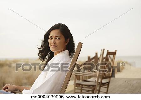 Stock Photograph - Asian woman sitting on deck at beach. Fotosearch