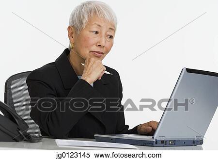 Stock Photography - Senior Asian businesswoman using laptop at desk. Fotosearch
