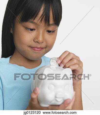 Stock Image - Young Asian girl putting coin in piggybank. Fotosearch