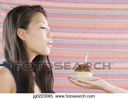 Stock Photography - Young Asian woman blowing out candle on cupcake. Fotosearch