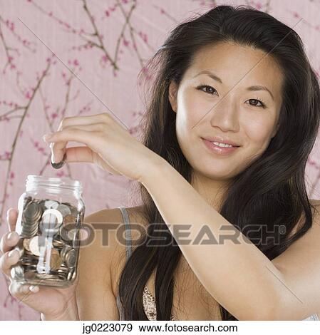 Young Asian woman putting coin in coin jar View Large Photo Image Stock Photo - Young Asian woman putting coin in coin jar. Fotosearch