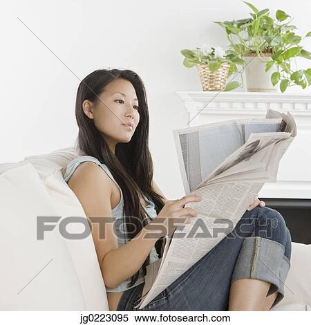 Stock Photography - Young Asian woman reading newspaper on sofa. Fotosearch