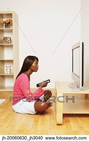 Stock Image - African woman sitting in front of television with remote control. Fotosearch
