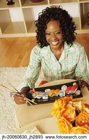 Stock Image - African woman sitting on floor eating sushi at home. Fotosearch