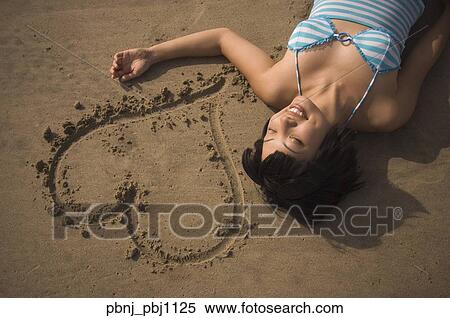 Stock Photography - Asian woman laying next to heart drawn in sand. Fotosearch