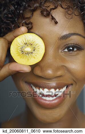 Stock Image - Close up of African woman holding slice of fruit in front of eye. Fotosearch