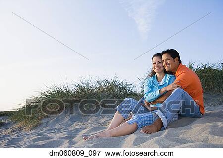 Stock Photo - Hispanic couple sitting on beach hugging. Fotosearch