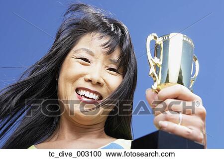 Stock Image - Low angle view of Asian woman holding trophy outdoors. Fotosearch