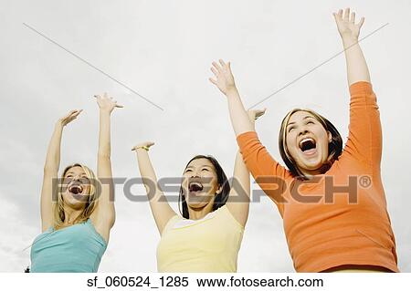 Stock Photography - Low angle view of three young woman laughing with arms raised outdoors. Fotosearch