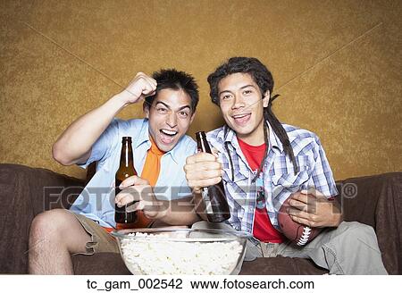 Two young men cheering on sofa with football View Large Photo Image Stock Image - Two young men cheering on sofa with football. Fotosearch